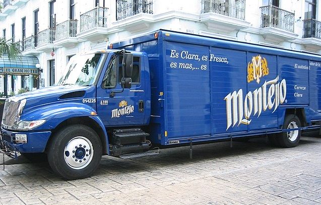 Prawo jazdy kategorii C. Na tle białego hotelu stoi niebieski samochód dostawczy z reklamą piwa Montejo.