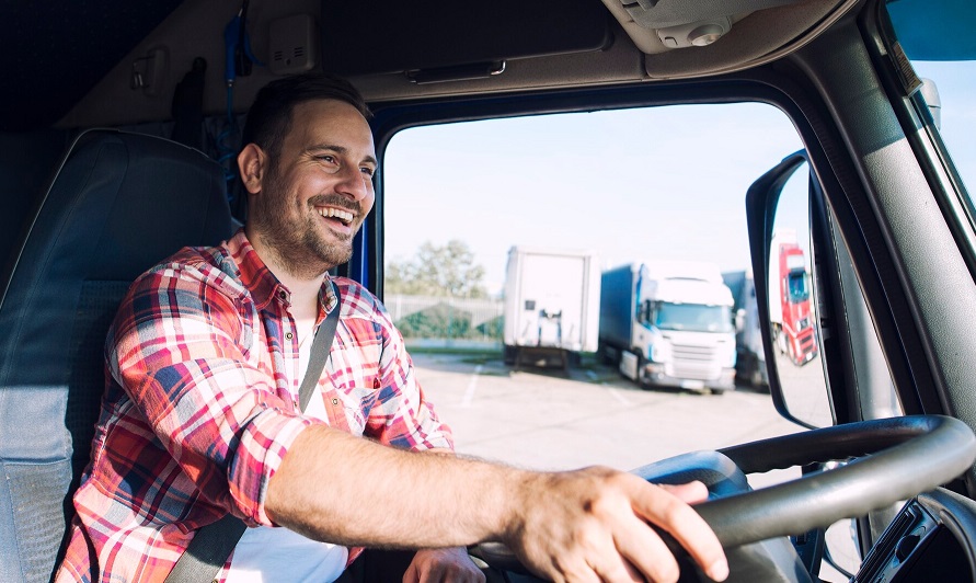 Karta kwalifikacji kierowcy. Młody, uśmiechnięty kierowca siedzi w kabinie ciężarówki. Twarz z lekkim zarostem, ubrany w kraciastą koszulę w odcieniach czerwieni.