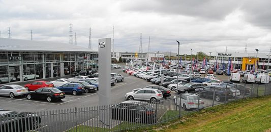 Wzrost cen samochodów używanych. Na pierwszym planie widoczny zielony zadbany trawnik. W oddali salon samochodowy Audi z samochodami nowymi i używanymi.