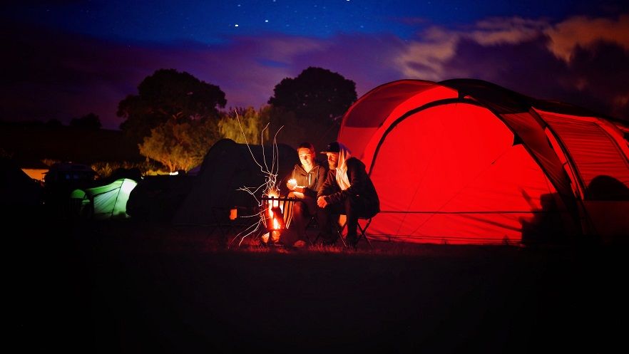 Bezpieczna majówka 2022. Leśny biwak nocą. Na pierwszym planie podświetlony czerwony namiot, przed nim przy ognisku siedzi para osób. W oddali inee podświetlone namioty. Biwak otoczony wysokimi drzewami nad nimi rozgwieżdżone niebo.