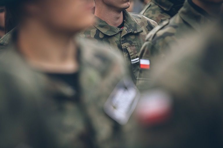 Kwalifikacja wojskowa 2022. Rozmyty obraz grupy żołnierzy w umundurowaniu polowym. Wyraźny jest tylko fragment rękawa z biało czerwoną naszywką.