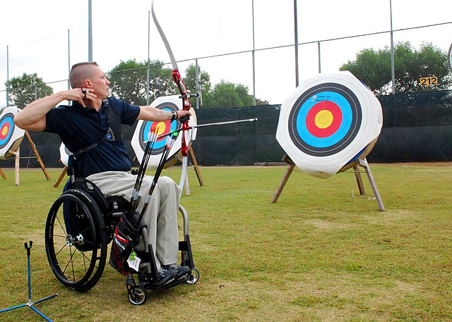 Na zdjęciu jest mężczyzna na wózku inwalidzkim strzelający z łuku do tarczy w zawodach. Łucznictwo osób z niepełnosprawnością.