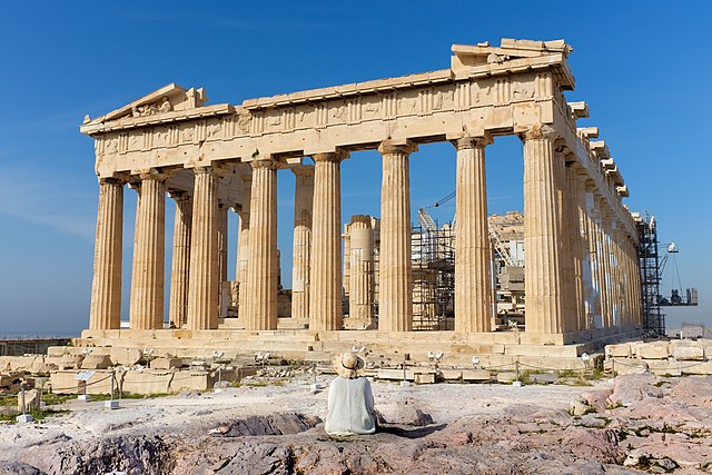 Prognozy na sezon wakacyjny 2022. Na tle lazurowego nieba wznoszą się pozostałości antycznej, greckiej budowli. Na skale przed jej frontem siedzi kobieta w słomkowym kapeluszu i podziwia kolumny pozostałe po świątyni.