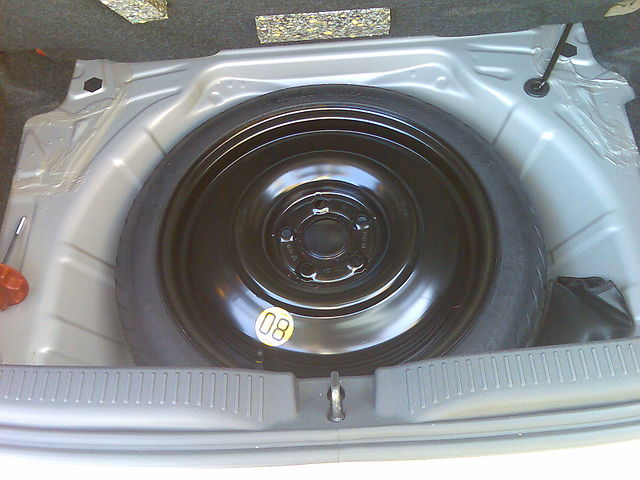 Koło zapasowe. Fotografia przedstawia wnękę w bagażniku srebrnej Toyoty Yaris. Wewnątrz leży zapasowe koło dojazdowe, które jest mniejsze i węższe od koła standardowego.