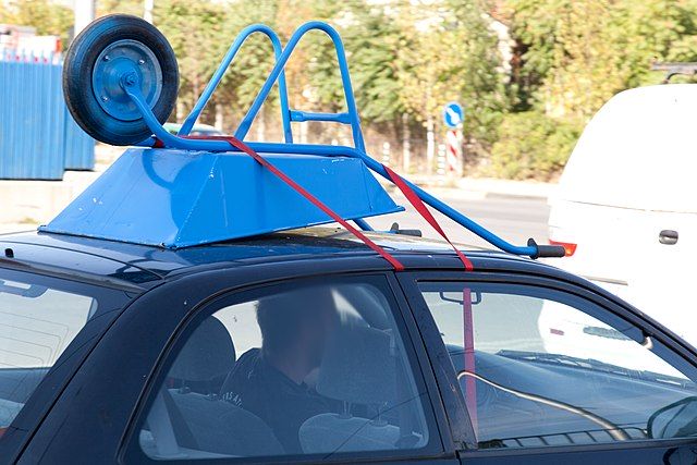 Bagażniki samochodowe. Na fotografii zbliżenie dachu małego samochodu miejskiego w kolorze granatowym. Kierowca przewozi na dachu taczkę, położoną bezpośrednio na niczym nie zabezpieczonym dachu.
