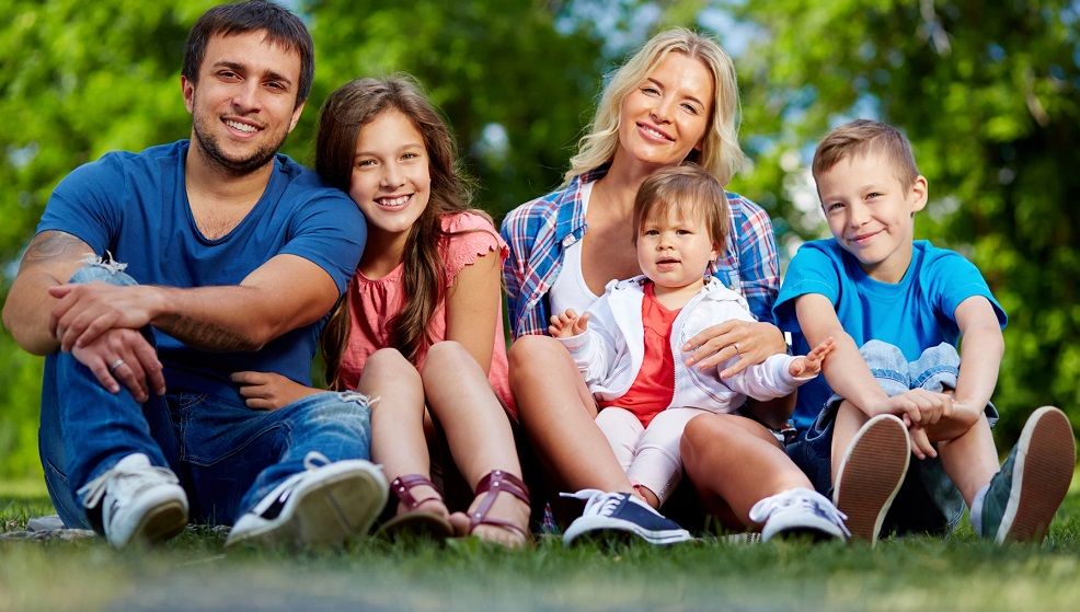 Bezwarunkowy Dochód Podstawowa 2022. Pięcioosobowa rodzina mama, tata oraz trójka dzieci siedzą uśmiechnięci na trawie. W tle zielone drzewa i krzewy.