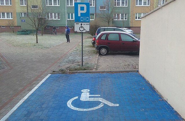 Karta parkingowa dla osób niepełnosprawnych. Na zdjęciu przykładowe puste miejsce parkingowe na blokowisku. Miejsce oznakowane niebieskim kolorem i znakiem niepełnosprawności.