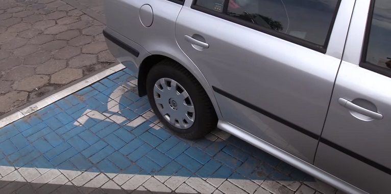 Karta parkingowa dla osób niepełnosprawnych. Na fotografii widoczny częściowo srebrny samochód kombi, zaparkowany na miejscu dla niepełnosprawnych.