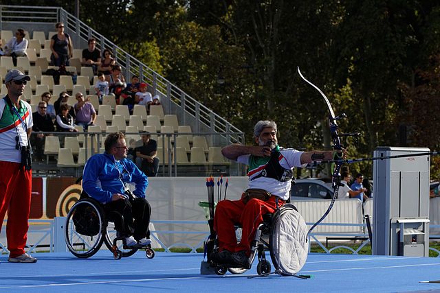 Na zdjęciu osoba na wózku inwalidzkim strzelająca z łuku do tarczy na zawodach. Łucznictwo osób z niepełnosprawnością.