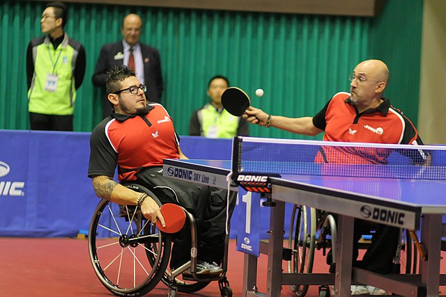 Na zdjęciu mężczyźni na wózkach inwalidzkich grający w deblu tenisa stołowego.