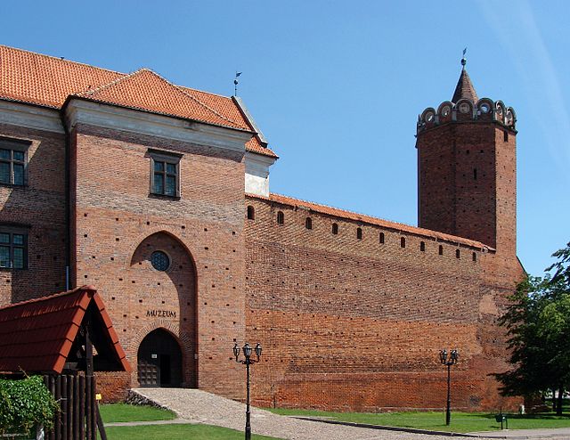 XXII Międzynarodowy Turniej Rycerski w Łęczycy. Widok na front zamku królewskiego w Łęczycy. Zamek zbudowany z cegły, widać po prawej narożną wieżę. Po lewej mieszkalna część zamku, wyższa od murów, kryta ceglastą dachówką. Na dole brama wjazdowa.