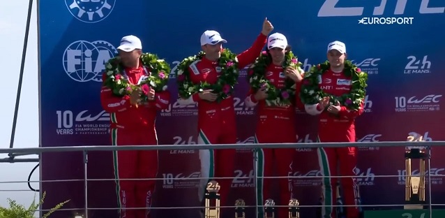 Zespół Kubicy drugi w Le Mans. Ta ilustracja jest zbliżeniem na zespół Prema Orlen Team stojący na podium. Czteroosobowa ekipa w czerwonych kombinezonach, na głowach białe czapeczki, a na szyjach zielone wieńce zwycięstwea.