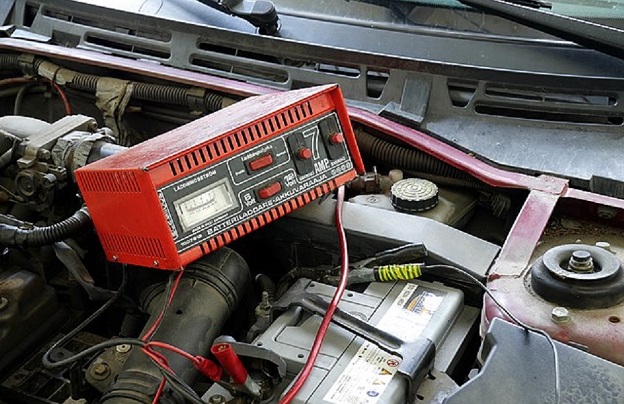 Akumulator rozruchowy - jaki wybrać. Na ilustracji widok na komorę silnika samochodu, maska w górze, a w centralnym punkcie czerwona skrzynka kablami i zegarami. To prostownik i właśnie odbywa się ładowanie akumulatora.
