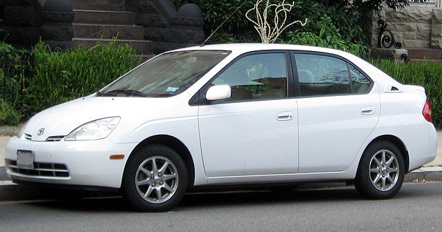 Samochód elektryczny czy hybrydowy. Na zdjęciu zaparkowana pod domem biała Toyota Prius pierwszej generacji z 1997 roku. Mały, biały samochód z małym bagażnikiem.