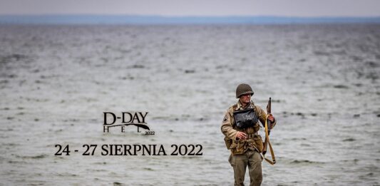 D-Day Hel 2022. Plakat w formie zdjęcia. Piaszczysta plaża, na krawędzi morskich fal biegnie rekonstruktor w mundurze amerykańskim 1944 roku z karabinem i w pełnym rynsztunku. Na tle morskich fal nazwa imprezy i data, czarnymi literami.