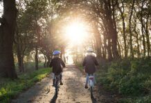 Bezpieczna jazda rowerem. Na zdjęciu w słoneczny dzień, dwójka dzieci jedzie na rowerach leśnym duktem. Oboje w kaskach jadą obok siebie.