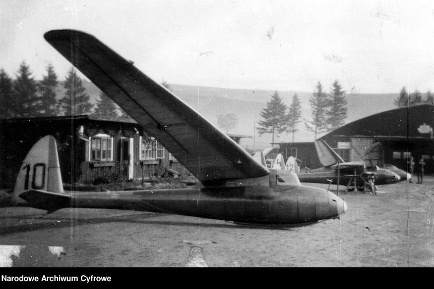III Zlot zabytkowych szybowców. Czarnobiałe zdjęcie z 1932 roku. Na nim w bocznym ujęciu szybowiec SG-28 przed hangarami. W oddali inne szybowce.