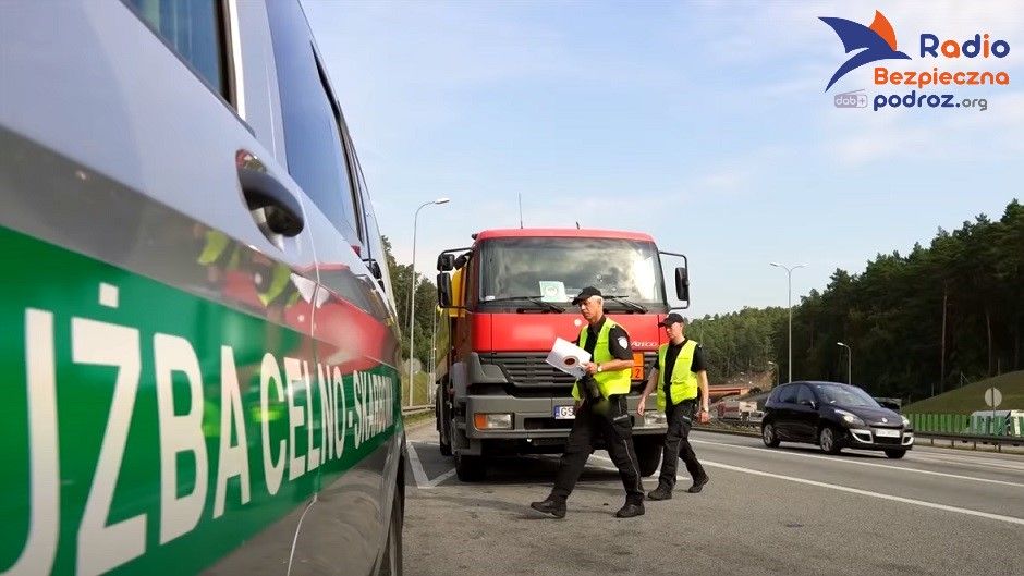 Kontrola drogowa - uprawnienia innych służb. Fotografia przedstawia dwóch funkcjonariuszy Służby Celno-Skarbowej idących z plikiem dokumentów przewozowych zatrzymanej ciężarówki. Akcja rozgrywa się na trasie szybkiego ruchu.
