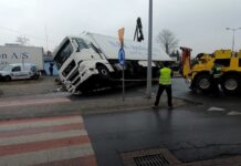 Całodobowa Pomoc Drogowa. Na zdjęciu, przewrócony biały tir z białą naczepą. Jest podnoszony przez ciężki samochód pomocy drogowej. Tir przewrócił się na prawy bok, prawdopodobnie wjechał na rondo ze zbyt dużą prędkością.