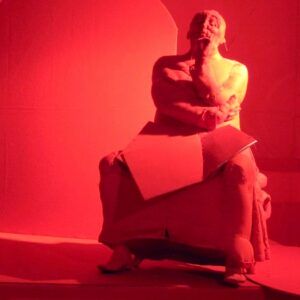 Wystawa rzeźby JAN KUCZ (1939-2021). Fotografia jednej z rzeźb na wystawie. Siedząca postać zamyślonego mężczyzny. Całość oświetlona silnym czerwonym światłem.