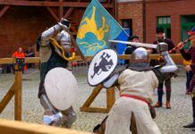 Turniej Rycerski Zamek w Ostródzie 2023. Na dziedzińcu zamkowym, dwaj rycerze walczą na miecze i tarcze w strojach z epoki.