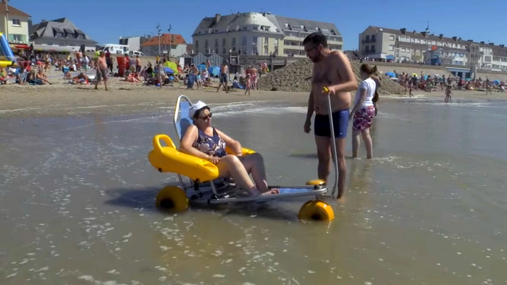 Urlop wypoczynkowy w 2024 roku, pomysły na wypoczynek osoby niepełnosprawnej. Na francuskiej plaży pan pomaga kobiecie niepełnosprawnej wejść do wody.
