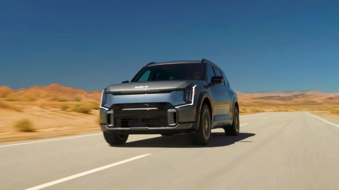 KIA-EV9-7-miejscowy-elektryk - Duży, srebrny, siedmioosobowy samochód elektryczny, jedzie asfaltową drogą przez amerykańską pustynię.