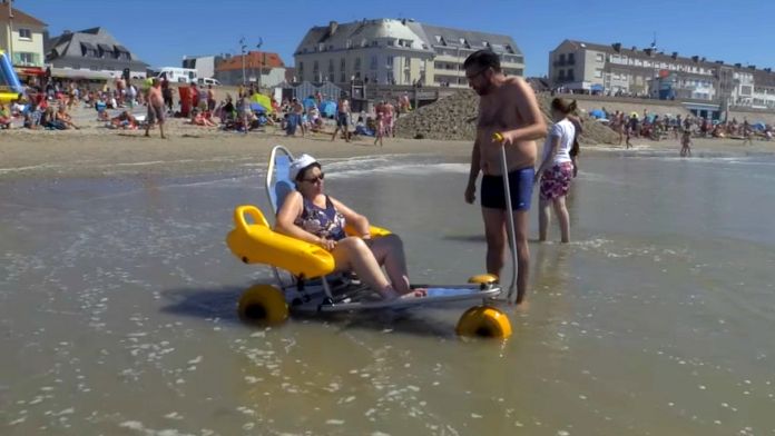 pomysły na wypoczynek osoby niepełnosprawnej. Na francuskiej plaży pan pomaga kobiecie niepełnosprawnej wejść do wody.