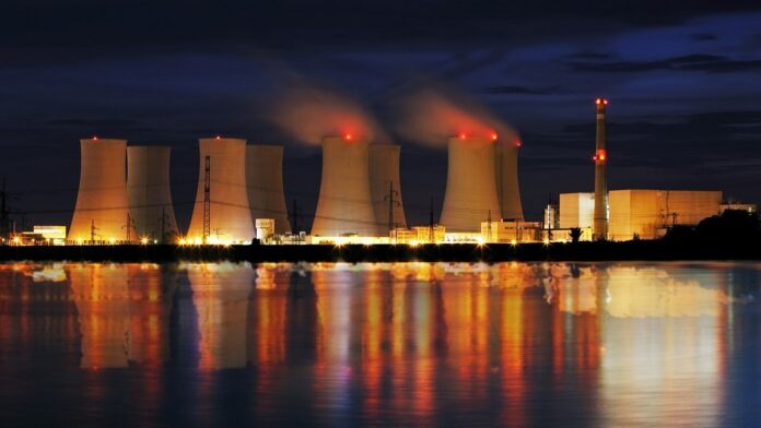 Budowa elektrowni atomowych w Polsce - nocne zdjęci elektrowni atomowej, mocno oświetlone kominy odbijają się na powierzchni wody pobliskiego zbiornika.