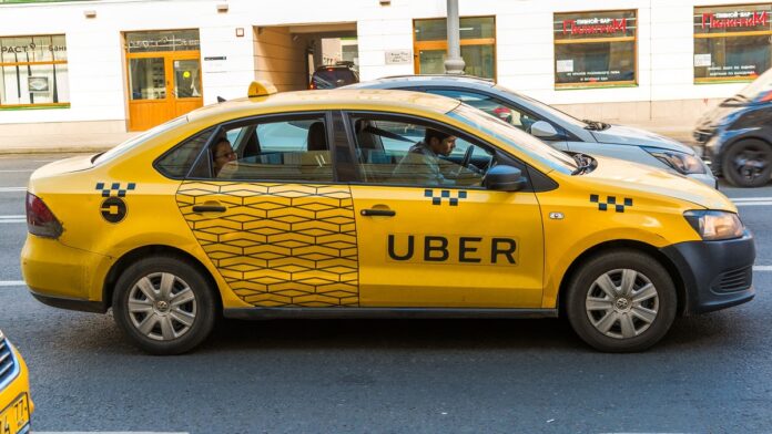 Firma Uber - żółta taksówka z czarnym napisem UBER na drzwiach. jedzie ulicą.