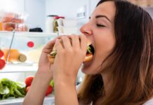 Świąteczne biesiady - młoda kobieta wpycha do ust ogromnego hamburgera. w tle otwarta lodówka
