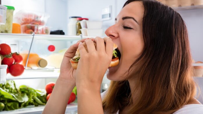 Świąteczne biesiady - młoda kobieta wpycha do ust ogromnego hamburgera. w tle otwarta lodówka