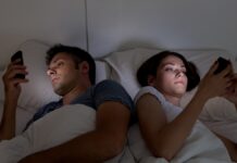 Uzależnienie od smartfona - dwoje młodych ludzi leży w łóżku. Odwróceni do siebie plecami, w dłoniach smartfony, ich twarze rozświetlone błękitną poświatą