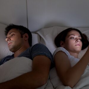 Uzależnienie od smartfona - dwoje młodych ludzi leży w łóżku. Odwróceni do siebie plecami, w dłoniach smartfony, ich twarze rozświetlone błękitną poświatą