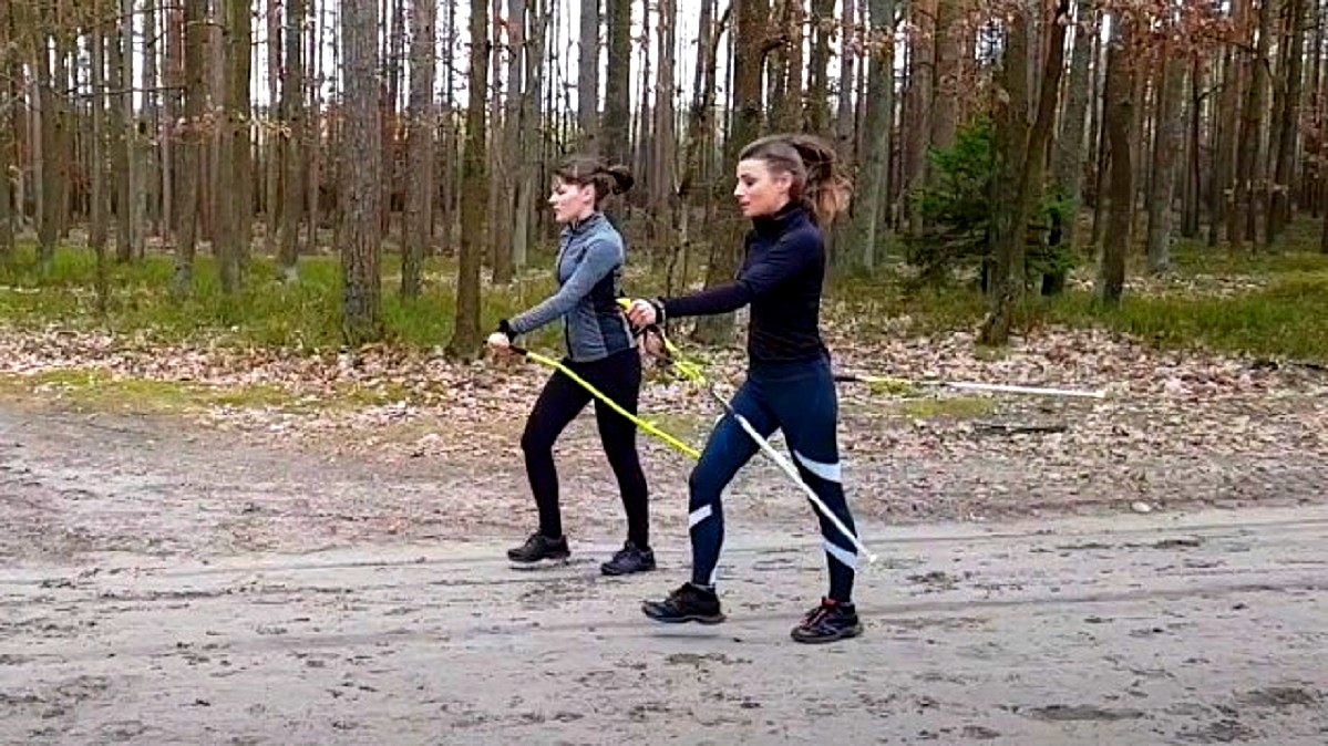 Dwie panie w dopasowanych strojach sportowych, uprawiają Nordic Walking podczas spaceru po lesie. Jesienny widok lasu, panie poruszają się po piaszczystym dukcie.