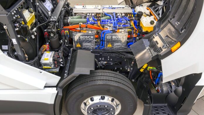 Firma BOSCH - zbliżenie na układ napędowy białego samochodu ciężarowego. Pod odchyloną do przodu kabiną kierowcy widać moduły zasilania ogniwami paliwowymi.
