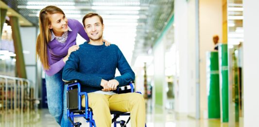 zmiany w przepisach dla niepełnosprawnych, uśmiechnięta kobieta i niepełnosprawny mężczyzna na wózku na korytarzu biurowca