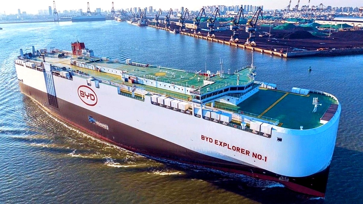 BYD chiński gigant - ogromny statek o nazwie BYDExplorer No1, wypływa z portu. Kadłub na górze biały, na dole brązowy, pokład zielony, kształt zaoblonego pudełka.