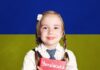 Nauka języka ukraińskiego w polskich szkołach - mała dziewczynka z warkoczykami stoi z ukraińskim podręcznikiem na tle ukraińskiej, niebiesko-żółtej flagi.