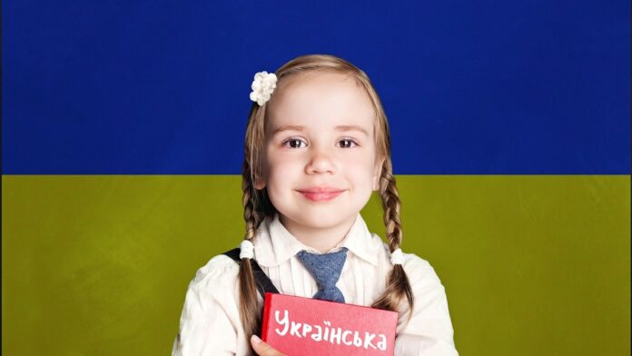 Nauka języka ukraińskiego w polskich szkołach - mała dziewczynka z warkoczykami stoi z ukraińskim podręcznikiem na tle ukraińskiej, niebiesko-żółtej flagi.
