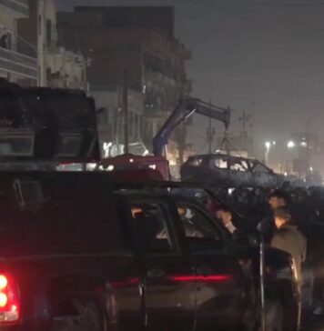 atak drona - nocny widok ulicy w Bagdadzie. Tłumy gapiów, policji i wojska. W oddali dźwig podnosi wrak samochodu zniszczonego przez drona.