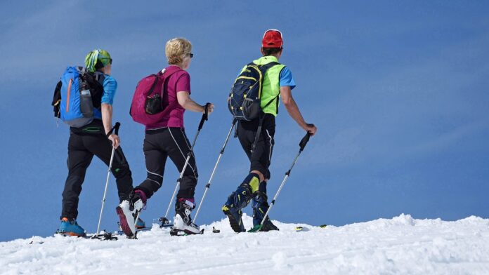 trasy narciarskie - trzech narciarzy w kolorowych strojach uprawia skitour. Idą po białym śniegu na tle błękitnego nieba.