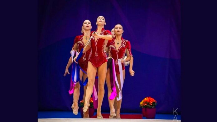 Złoto Pucharu Świata - zespół polskich gimnastyczek w czerwonych strojach, z kolorowymi wstążkami w rękach wychodzi na scenę. Granatowe tło.