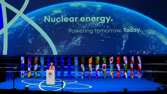 energetyka jądrowa - widok z daleka na scenę ze stojącymi flagami 11 państw UE. Po lewej z mównicy przemawia przewodnicząca w łososiowym żakiecie. Na ogromnym telebimie widok Ziemi z kosmosu oraz napis z tytułem konferencji.