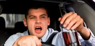 pijany ukrainiec - zbliżenie na młodego, agresywnego kierowcę. Prawa ręka na kierownicy, w uniesionej lewej upita butelka alkoholu.