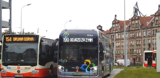wodorowy NesoBus - na pętli autobusowej obok biało-czerwonego autobusu gdańskiej komunikacji, stoi nowoczesny, srebrny autobus z licznymi wielobarwnymi aplikacjami oraz hasłami zachwalającymi napęd wodorowy.