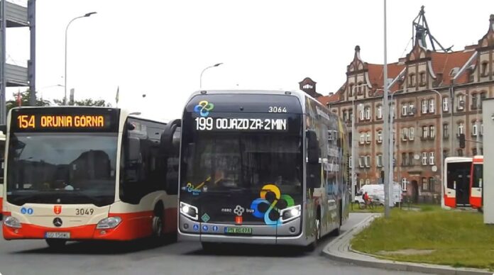 wodorowy NesoBus - na pętli autobusowej obok biało-czerwonego autobusu gdańskiej komunikacji, stoi nowoczesny, srebrny autobus z licznymi wielobarwnymi aplikacjami oraz hasłami zachwalającymi napęd wodorowy.