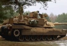Czołgi Abrams M1A1 - w sosnowym lesie stoi czołg widziany z prawego profilu. Malowanie maskujące brązowo-zielone. Widać żołnierzy z obsługi czołgu.