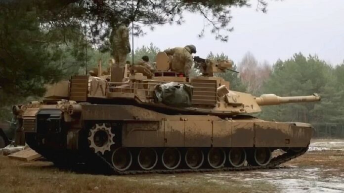 Czołgi Abrams M1A1 - w sosnowym lesie stoi czołg widziany z prawego profilu. Malowanie maskujące brązowo-zielone. Widać żołnierzy z obsługi czołgu.