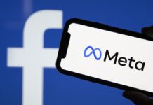 Meta (dawny Facebook) - zbliżenie na ekran smartfona. Na białym tle duże logo Mety. N drugim planie niebieska ściana z białym logo facebooka.